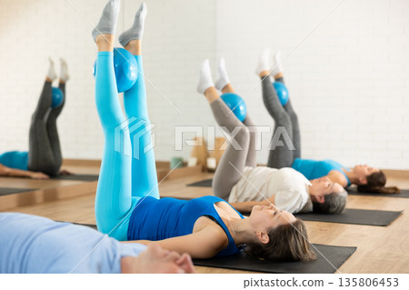 People of different ages pressing core and clutching mini Pilates balls between legs during Pilates training together 135806453