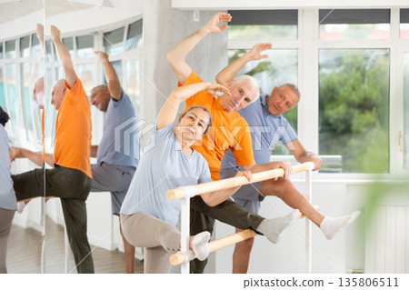 Dedicated mature females and males ballet dancer performing leg stretch at barre with concentration and grace in choreography studio during group rehearsal 135806511