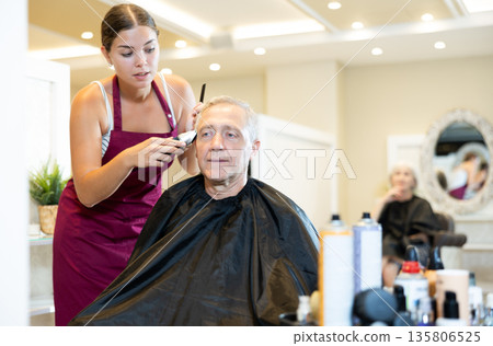 Young female hairdresser cutting hair of elderly client with clipper in salon Young female hairdresser cutting hair of elderly client with clipper in salon 135806525