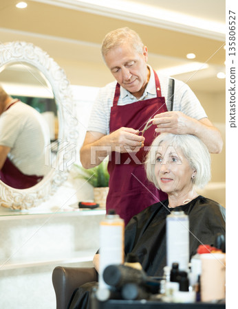 Professional male hairdresser cuts hair of elderly woman at beauty salon 135806527
