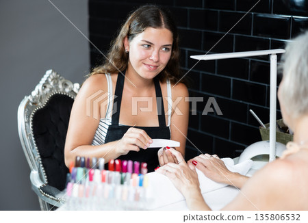 Girl manicurist works with unrecognizable pensioner client, files nails and performs hand care 135806532
