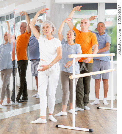 Experienced mature female ballet dancer, dressed in sportswear and group of women and men engaged in classical ballet performs exercise standing in 3rd position near ballet barre in dance hall 135806534