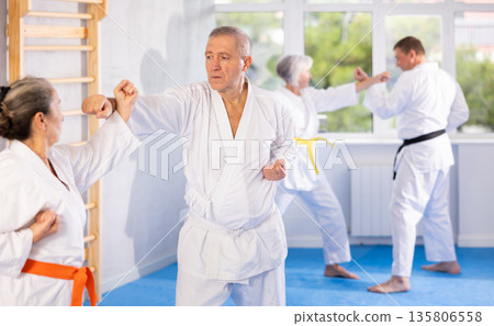 Old man attendee of karate classes fighting with his opponent in sports hall Old man attendee of karate classes fighting with his opponent in sports hall 135806558