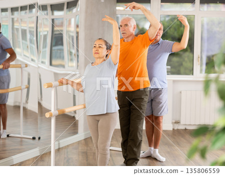 Ordinary elderly people stand in third position near ballet barre during group training in dance studio Ordinary elderly people stand in third position near ballet barre during group training in dance studio 135806559