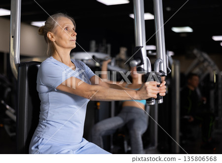 Elderly female training trains muscles of arms and press on simulator in gym 135806568