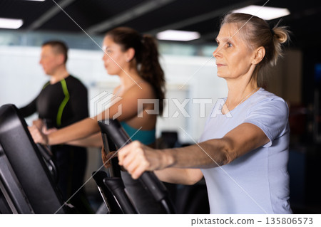 Mature woman exercising on an elliptical trainer in gym Mature woman exercising on an elliptical trainer in gym 135806573