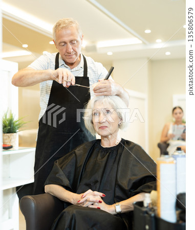 Experienced senior man master makes performs haircut for mature woman with scissors 135806579