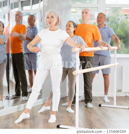 Active mature woman practicing battement tendu holding by ballet barre together with others 135806584