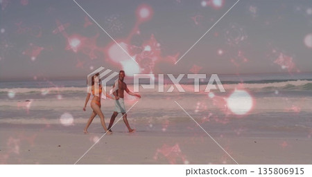 Strolling couple in bikini and swim trunks moving along beach shoreline, with pink star overlay 135806915