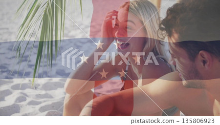 Laughing couple wearing bikini top and swim trunks on white sand under palm frond, tropical beach Laughing couple wearing bikini top and swim trunks on white sand under palm frond, tropical beach 135806923