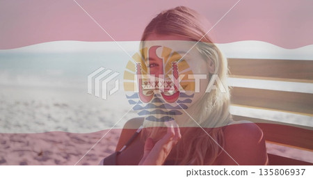 Relaxing woman wearing red bikini sitting on wooden lounge chair at beach, with emblem flag overlay Relaxing woman wearing red bikini sitting on wooden lounge chair at beach, with emblem flag overlay 135806937