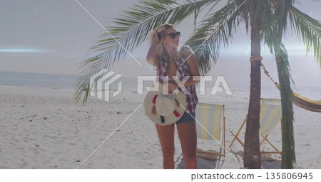 Holding straw hat and touching hair, woman wearing sunglasses posing on sandy beach by hammock 135806945