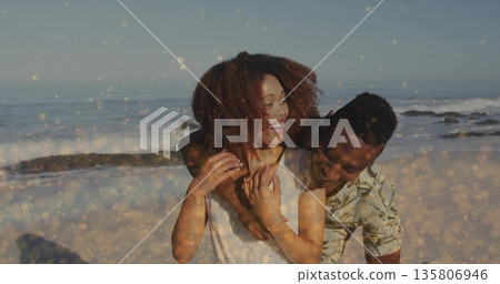 Hugging laughing couple wearing sleeveless top and tropical shirt at sandy beach, sparkling waves 135806946