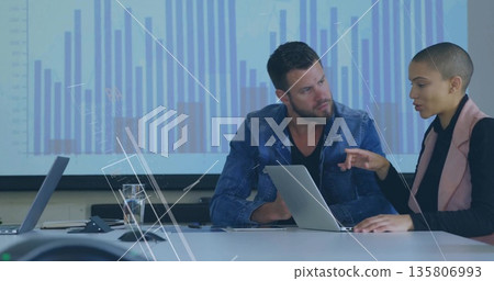 Discussing insights, two colleagues reviewing bar chart in meeting room, with laptops and phone 135806993