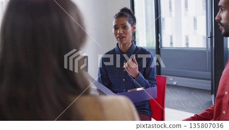Holding folder and leading discussion, woman in denim shirt speaking at office windows Holding folder and leading discussion, woman in denim shirt speaking at office windows 135807036