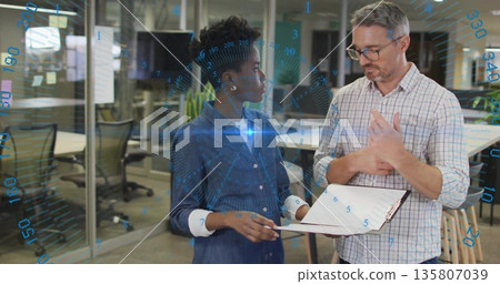 Coworkers discussing digital interface overlay near glass-wall office desks, holding clipboard Coworkers discussing digital interface overlay near glass-wall office desks, holding clipboard 135807039