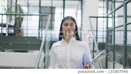 Walking businesswoman carrying color-tipped folders through office atrium, showing digital network 135807040