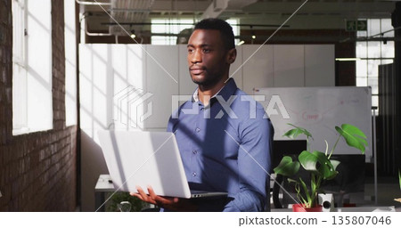 Standing office worker holding silver laptop in open-plan office, with whiteboard and plant Standing office worker holding silver laptop in open-plan office, with whiteboard and plant 135807046