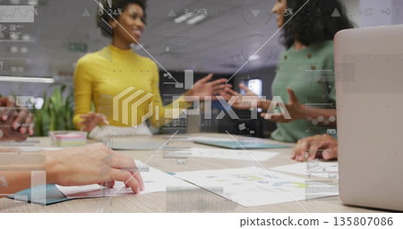 Gesturing colleagues wearing mustard and green sweaters at meeting table with charts, silver laptop 135807086