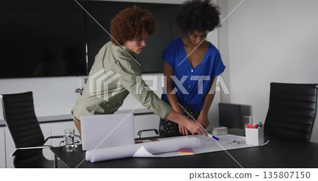 Leaning over rolled-out blueprint two colleagues collaborating at table, with laptop, sticky notes 135807150
