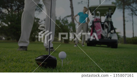 Positioning driver club head behind golf ball on tee at tee box, with golf cart, bags 135807161