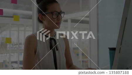 Working Asian woman in sleeveless blouse in open-plan office, with monitor, sticky notes, teal mug Working Asian woman in sleeveless blouse in open-plan office, with monitor, sticky notes, teal mug 135807188