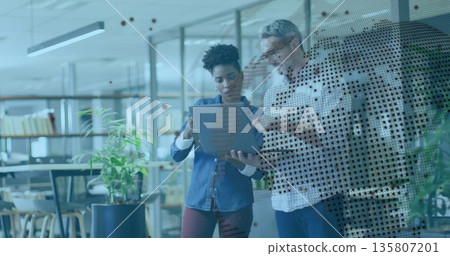 Reviewing laptop two coworkers standing behind glass partition in office, with bookshelves, plants Reviewing laptop two coworkers standing behind glass partition in office, with bookshelves, plants 135807201