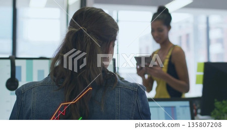 Denim-jacketed woman analyzing charts on monitors in open office, with colleague glancing at tablet Denim-jacketed woman analyzing charts on monitors in open office, with colleague glancing at tablet 135807208