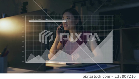 Chinese woman wearing burgundy top holding smartphone and using computer in office, with papers Chinese woman wearing burgundy top holding smartphone and using computer in office, with papers 135807216