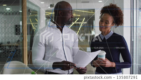 Mature man and woman discussing reports in office lobby, with coffee cup, glass partition Mature man and woman discussing reports in office lobby, with coffee cup, glass partition 135807291