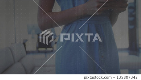 Standing woman in blue dress holding smartphone in lounge area, with sofa, table, chairs and lamp Standing woman in blue dress holding smartphone in lounge area, with sofa, table, chairs and lamp 135807342