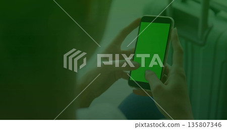 Holding smartphone green screen while woman traveler sitting at terminal seating, carrying suitcase Holding smartphone green screen while woman traveler sitting at terminal seating, carrying suitcase 135807346