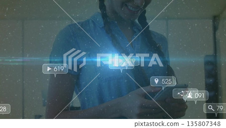 Smiling Hispanic woman wearing denim shirt holding smartphone at home, floating social metric icons 135807348