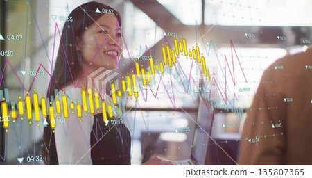 Holding laptop, Chinese businesswoman reviewing digital stock charts at modern office with coworker 135807365