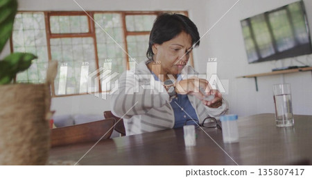 Opening Indian senior woman taking pill from bottle at home dining table, with glass of water 135807417