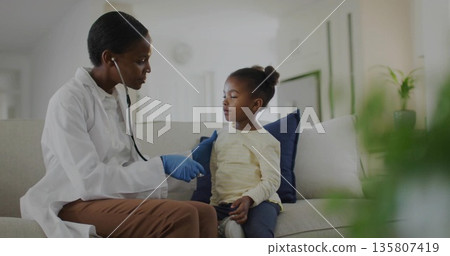 Doctor wearing lab coat and blue gloves using stethoscope on girl on sofa in living room 135807419