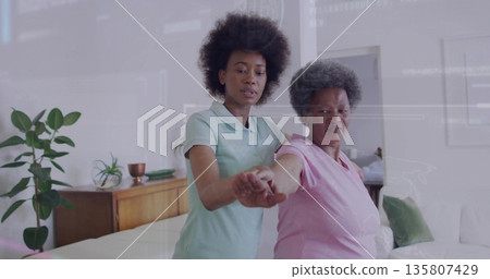 Assisting senior woman with guided arm stretch in living room, with potted plant on sideboard 135807429