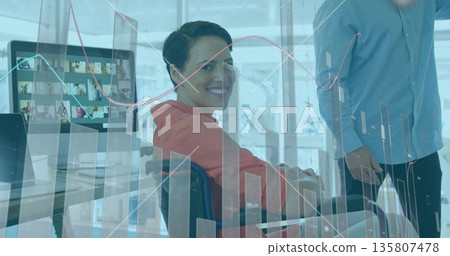 Smiling orange-top and blue-shirt colleagues discussing data at office desk with monitor and charts 135807478