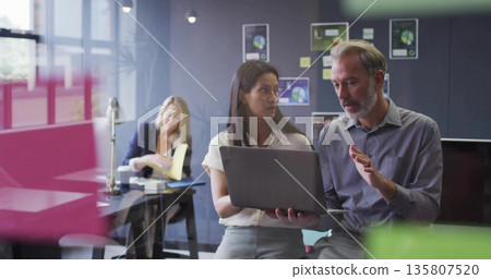 Discussing business colleagues reviewing laptop in open office, with charts and sticky notes Discussing business colleagues reviewing laptop in open office, with charts and sticky notes 135807520