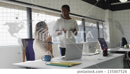 Discussing business-casual colleagues using tablet and laptop at desk in office, with digital map Discussing business-casual colleagues using tablet and laptop at desk in office, with digital map 135807526