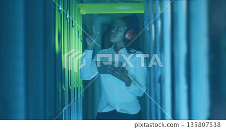 Inspecting female IT technician adjusting server module in data center aisle with tablet, earmuffs 135807538