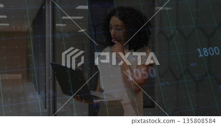 Holding laptop, woman in blouse analyzing chart overlay with gridlines in office, glass partitions 135808584