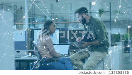 Collaborating software professionals reviewing code at open-plan office desk, with dual monitors 135808586