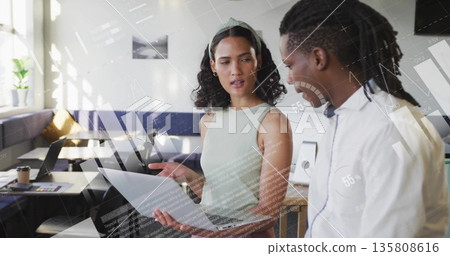 Standing colleagues in casual business attire discussing document in office, with laptop and coffee Standing colleagues in casual business attire discussing document in office, with laptop and coffee 135808616
