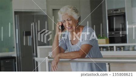Leaning senior woman holding smartphone to ear in modern kitchen, with railing and potted plant 135808621