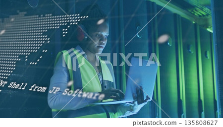 Tech wearing hi-vis vest, red earmuffs reading laptop code overlay in data center before racks Tech wearing hi-vis vest, red earmuffs reading laptop code overlay in data center before racks 135808627