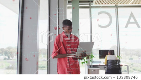 Man in red shirt holding laptop reviewing code in office with glass walls, with desktop monitor Man in red shirt holding laptop reviewing code in office with glass walls, with desktop monitor 135809746