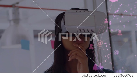 Standing Asian woman wearing VR headset thinking in office with sticky notes, showing network icons 135809765