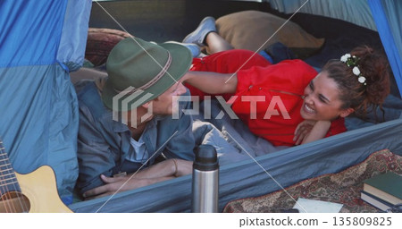 Lying couple wearing fedora red zip-up top inside sleeping bag lined tent, with guitar and books Lying couple wearing fedora red zip-up top inside sleeping bag lined tent, with guitar and books 135809825