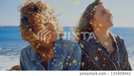 Gazing two women wearing denim jacket, patterned button-down shirt standing on rocky shore by ocean 135809842